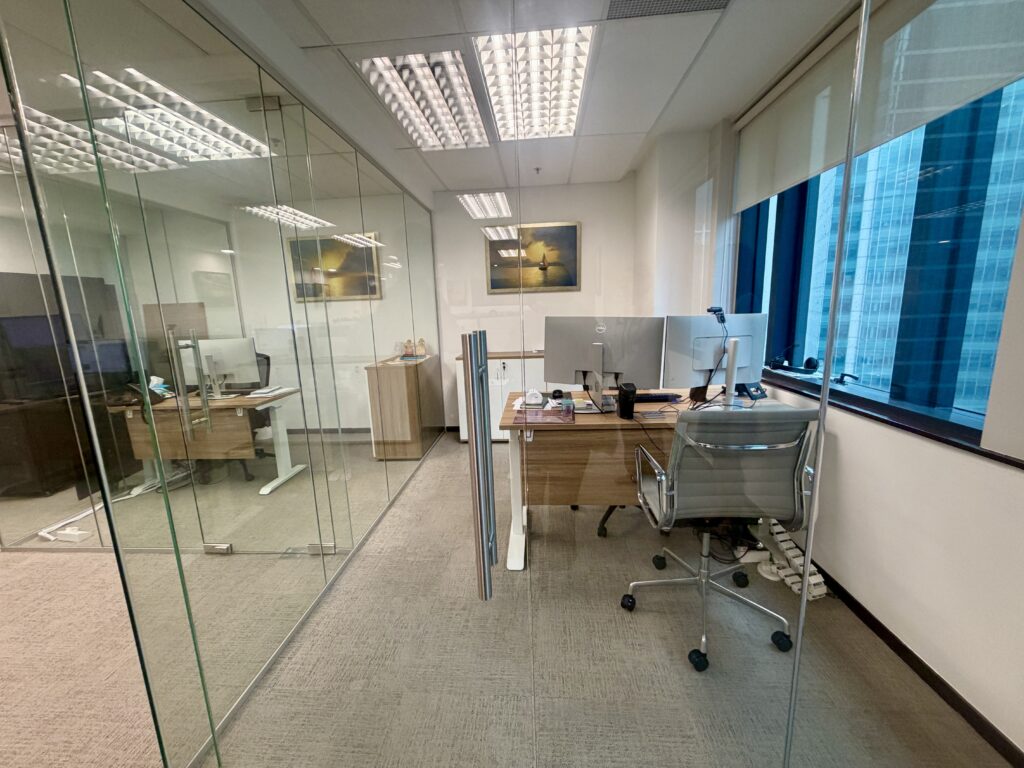 A modern office with two glass-walled workstations, each featuring a wooden desk, dual monitors, and an ergonomic chair.