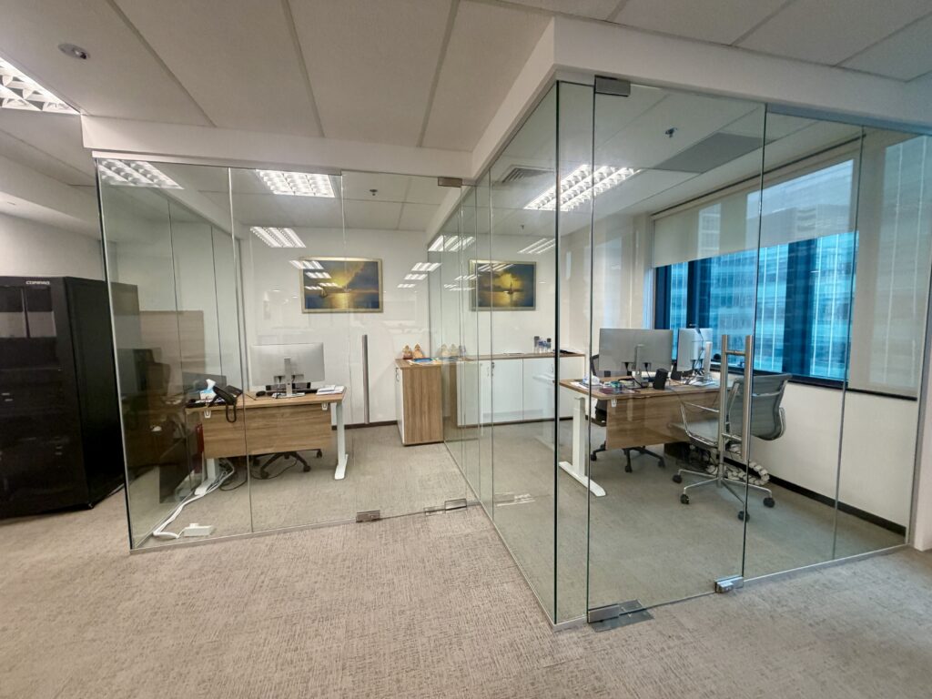Two adjacent glass-walled office cubicles, each containing a wooden desk, computer, and office chair.