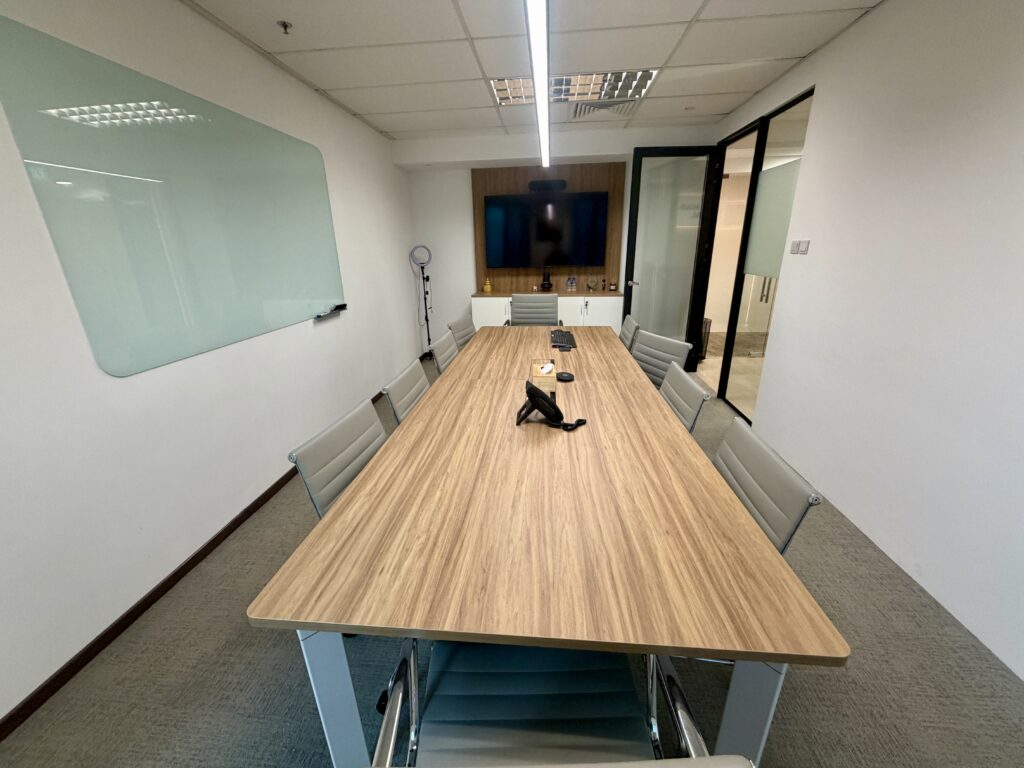 A conference room with a long wooden table, chairs, a wall-mounted monitor, and a glass whiteboard.