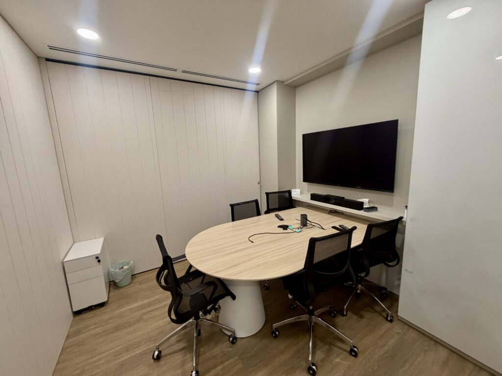 Small meeting room with an oval wooden table, four black office chairs, and a wall-mounted television.