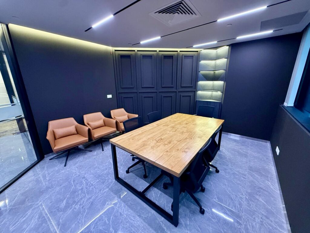 A conference room with a wooden table, four black office chairs, and three tan lounge chairs against a dark wall with recessed shelving.