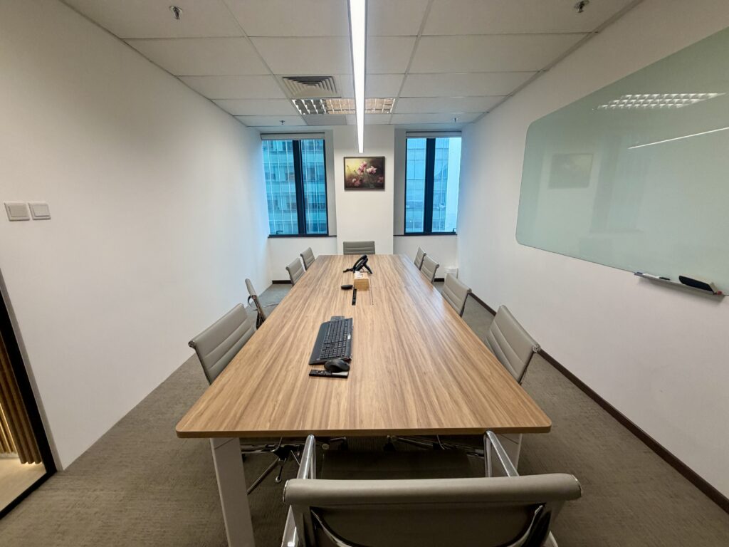 A wood-topped conference table surrounded by grey office chairs in a meeting room with a glass whiteboard.