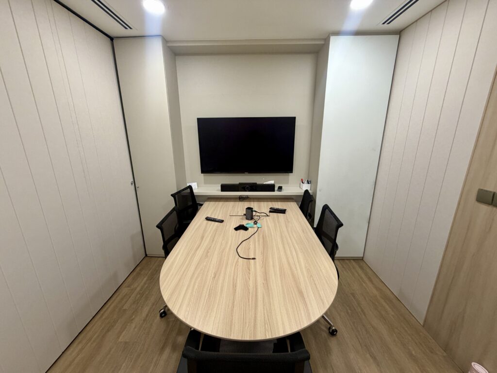 A small meeting room with an oval wooden table, four chairs, and a wall-mounted television.