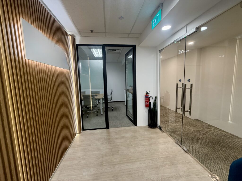 Office corridor with a wood-slatted wall, a glass-walled meeting room, and a glass-panelled entrance with an exit sign overhead.
