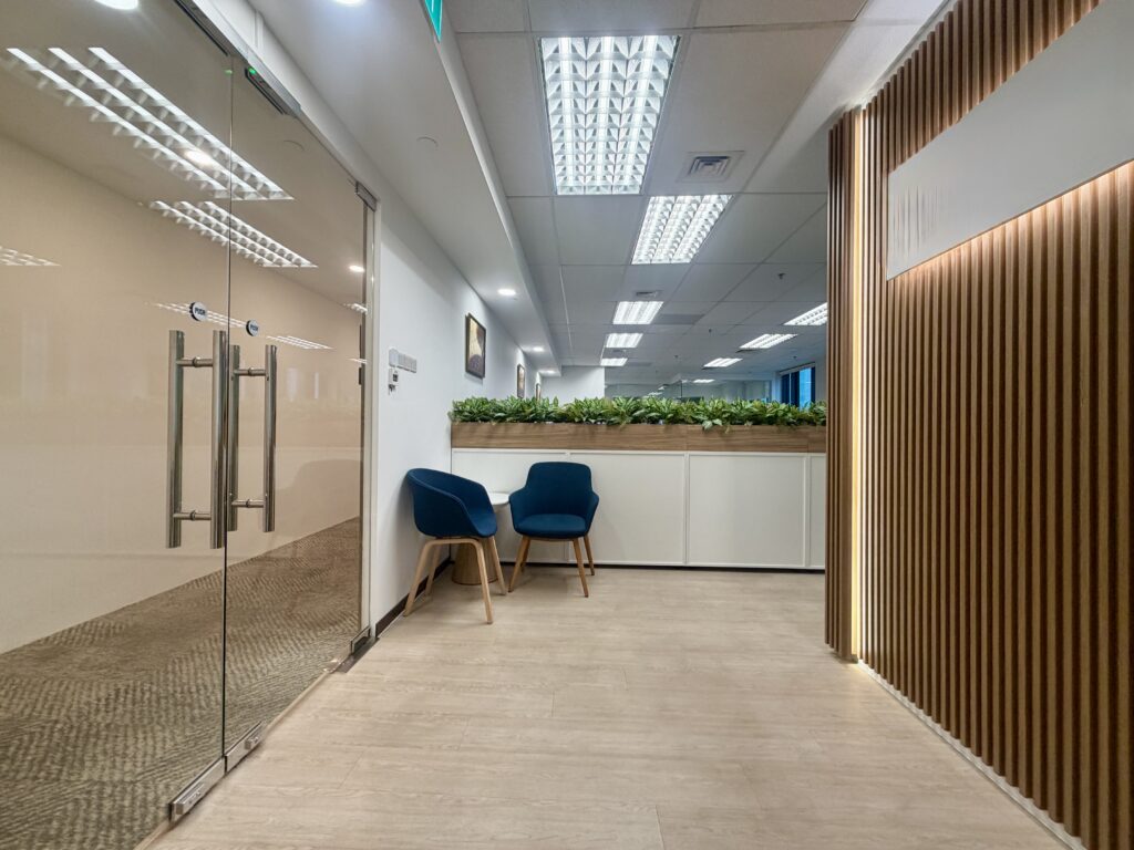 A modern office waiting area featuring two blue chairs next to a glass-walled meeting room and a wood-panelled wall.