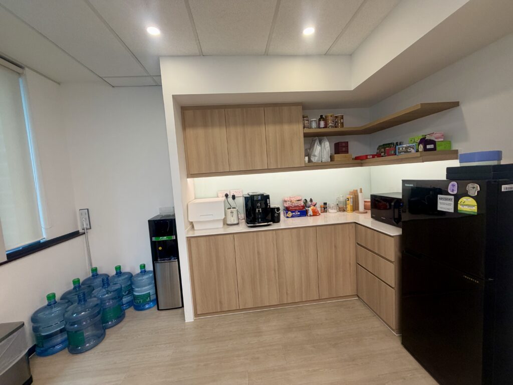 Office kitchenette with light wood cupboards, a black coffee machine, snacks, a water dispenser, several large water bottles, and a black fridge.