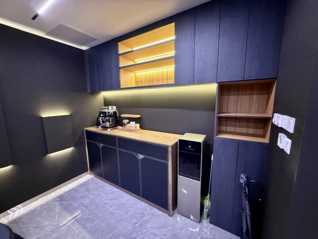 Kitchenette with dark cupboards, wood-tone open shelving, a coffee machine, and a floor-standing water dispenser on a grey tiled floor.