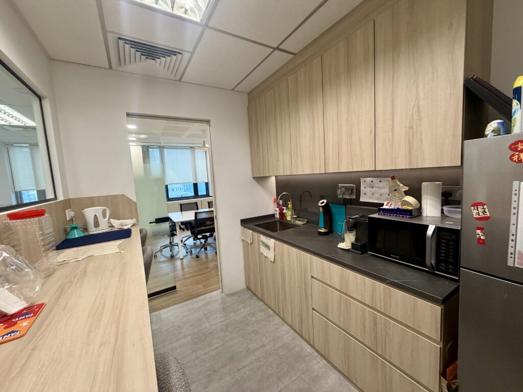 A small office kitchenette with wood-toned cupboards, a black worktop, sink, microwave, and a doorway leading into an office workspace.