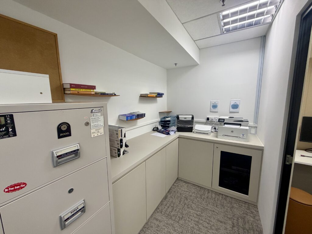 A small office nook with a beige filing cabinet, L-shaped worktops, a printer, and floating shelves holding a few books.