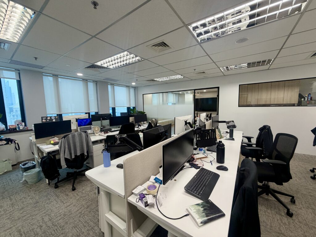 An open-plan office space featuring rows of white desks equipped with computer monitors, keyboards, and office chairs.