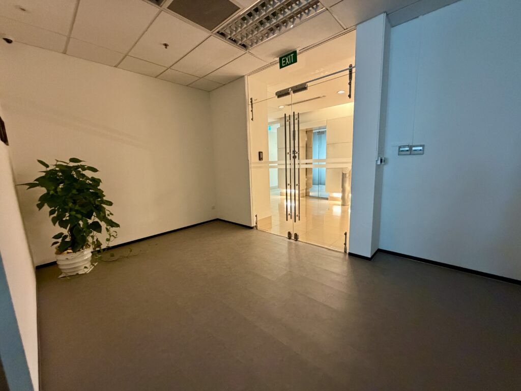 An empty office corridor with a potted plant in the corner, leading to a glass door with a green “EXIT” sign above it.