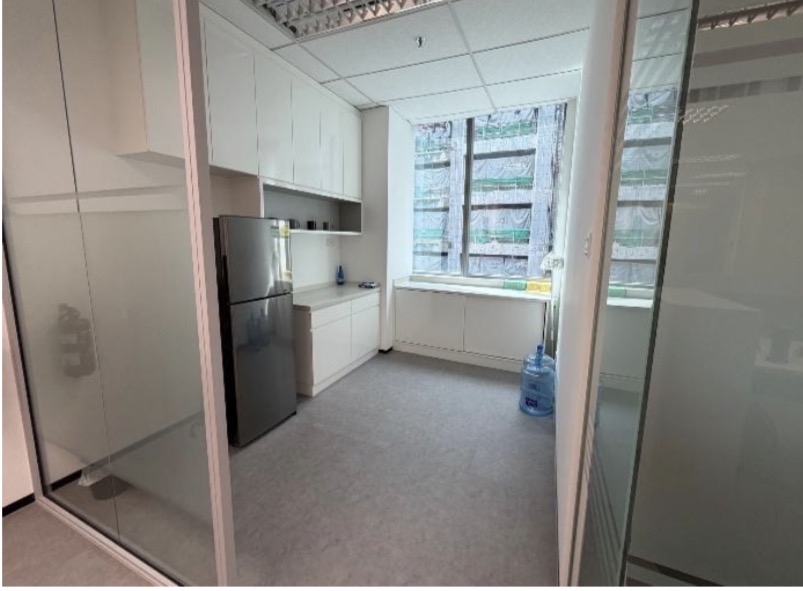 Small office kitchenette featuring a fridge, white cupboards, and a large window.