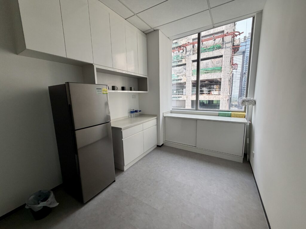 A kitchenette with a stainless steel fridge and white cupboards, next to a large window overlooking a building under construction.