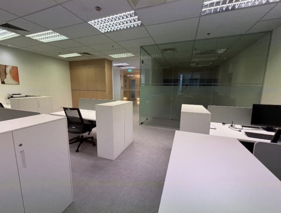 An open-plan office with white workstations, storage cupboards, and a glass-walled meeting room.