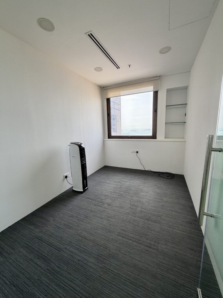An empty office room with grey carpet, a window, built-in shelving, and a portable air cooler.