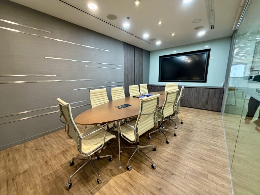 Oval conference table with eight cream-coloured rolling chairs in a room featuring a large wall-mounted monitor.