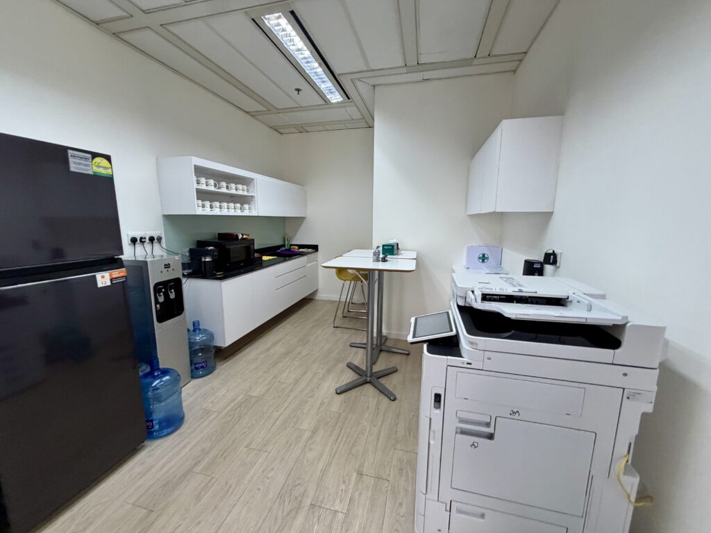 An office pantry featuring a fridge, water dispenser, cupboards with mugs, a small table, and a large printer.