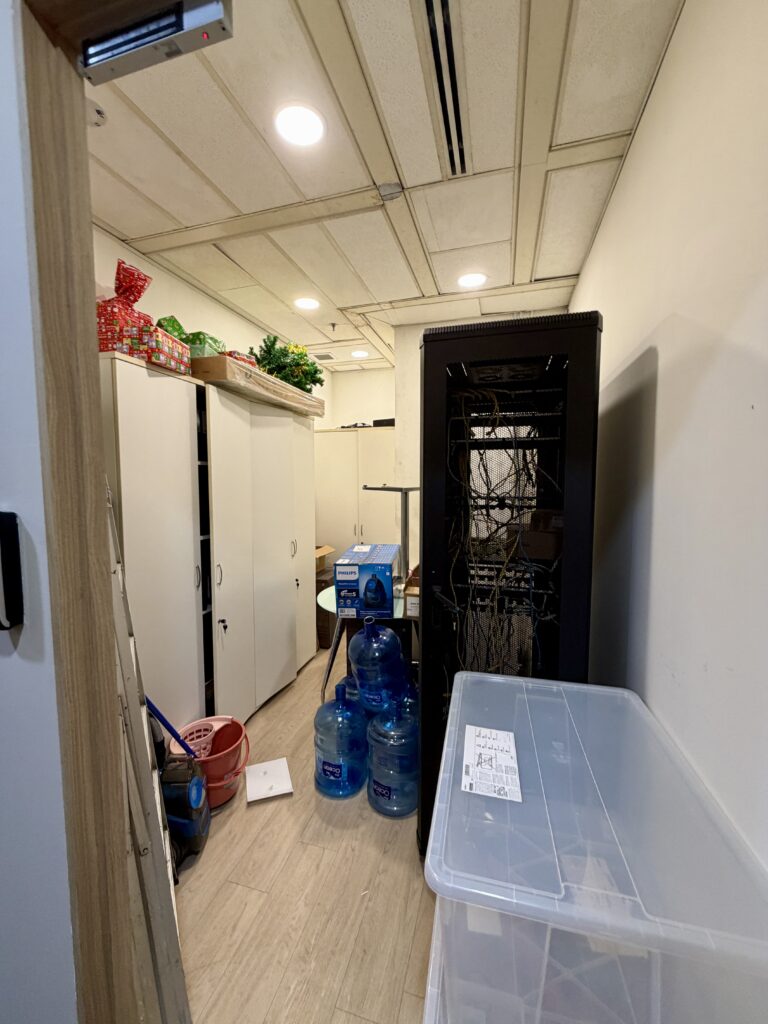 A narrow utility room containing white storage cupboards, a tall server rack, stacks of water containers, and a translucent storage box.