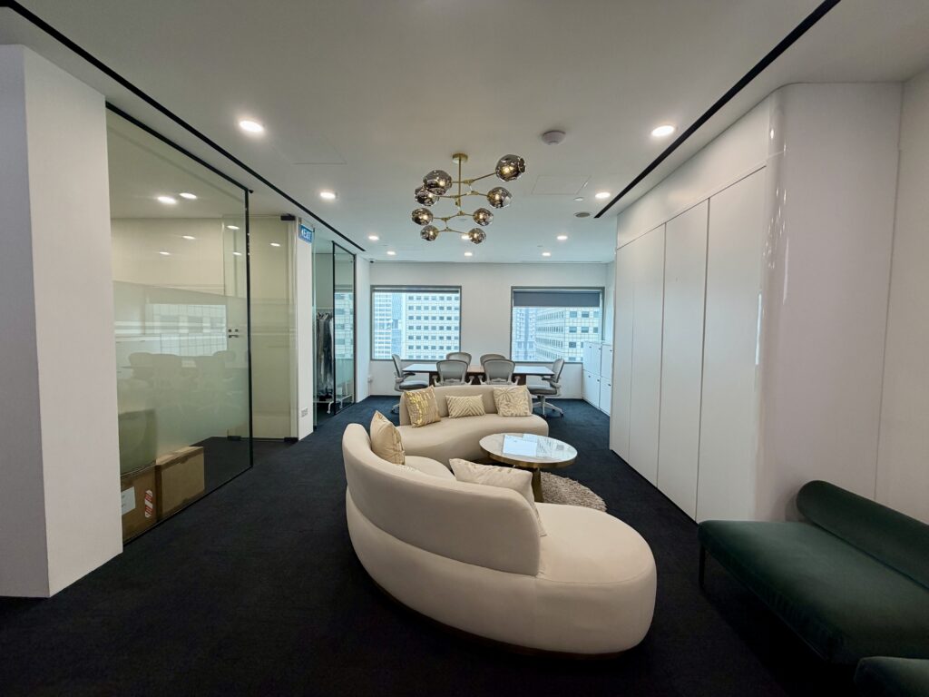 A modern office lounge with a curved white sofa, coffee table, and a glass-walled meeting room in the background.