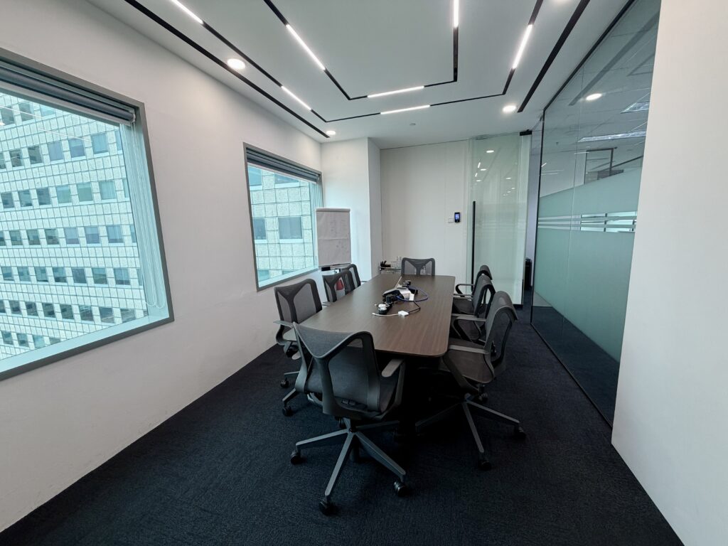 A conference room with a rectangular table, six swivel chairs, a whiteboard, and large windows overlooking city buildings.