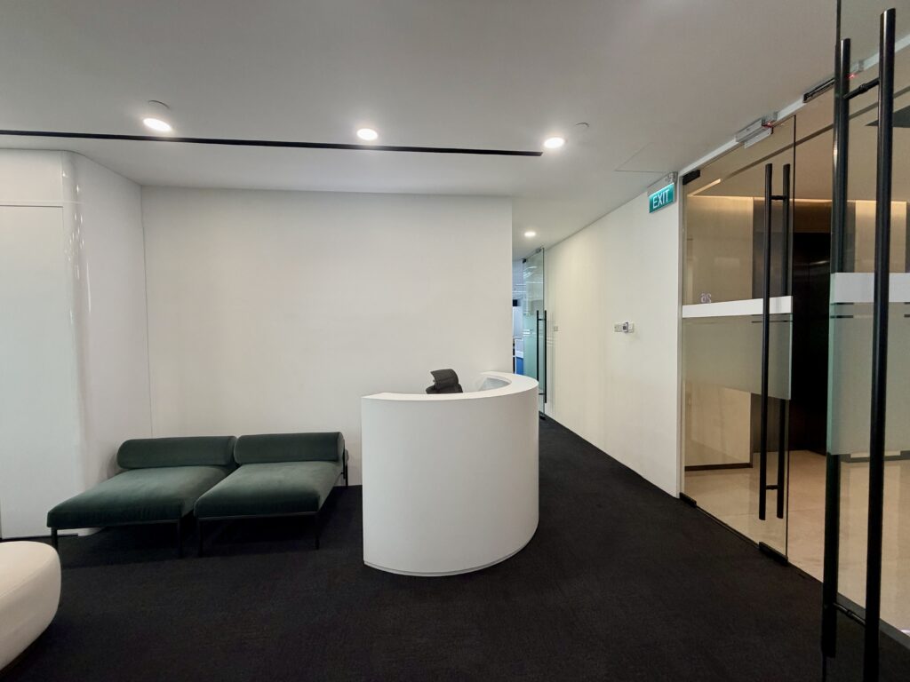 Modern office reception area with a curved white desk, a dark green bench, and glass doors.
