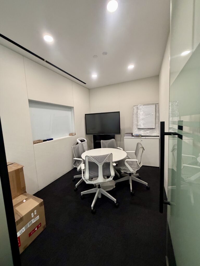 Small meeting room with a round white table, four office chairs, a wall-mounted television, and a whiteboard.