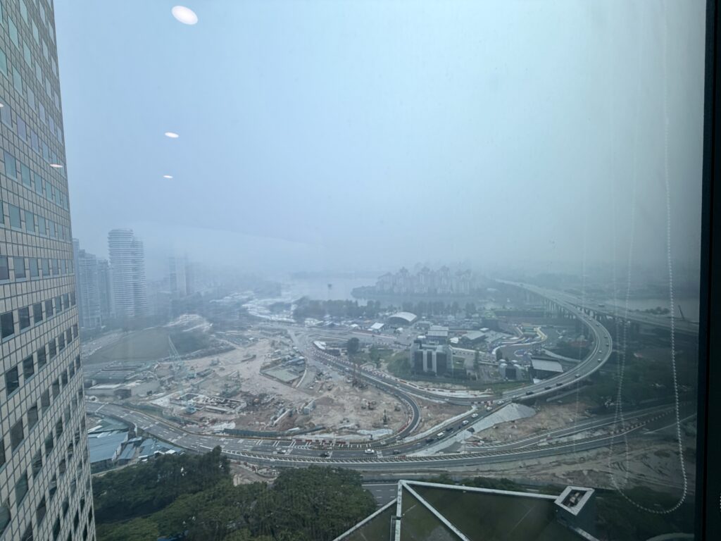 High-angle view through a window showing a cityscape with motorway interchanges, construction sites, and buildings under a hazy, overcast sky.